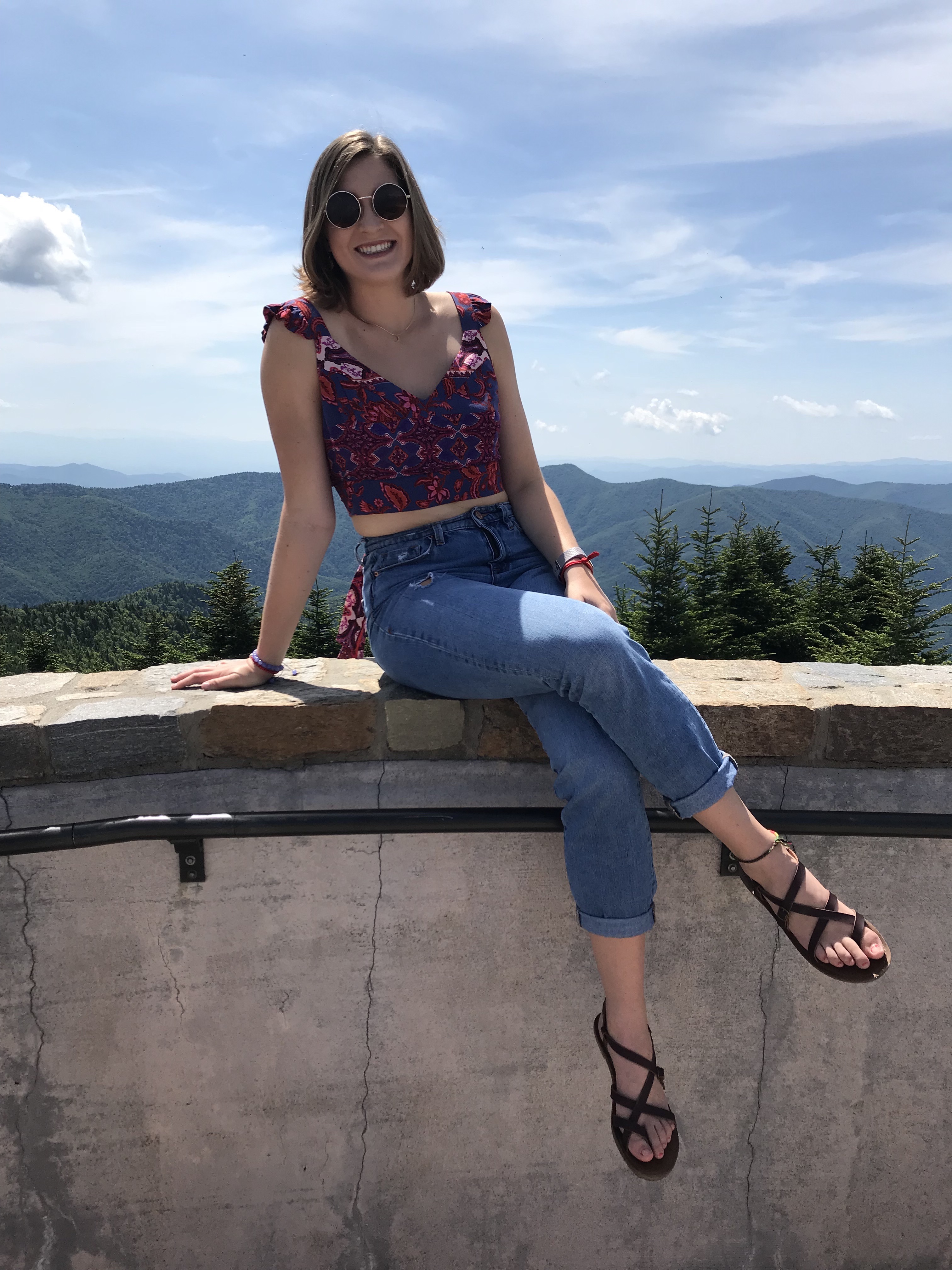 Woman sitting on edge of railing with mountains and trees in background.